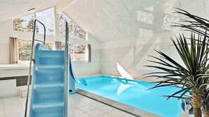 An indoor swimming pool with a blue slide, a potted plant, and a view of trees through the windows. - 5 Star Holiday Home in Liseleje (Liseleje)