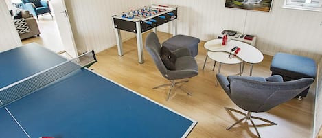 A room with a ping pong table, a foosball table, two grey armchairs, a small white table with a red bottle, and a framed picture on the wall.
