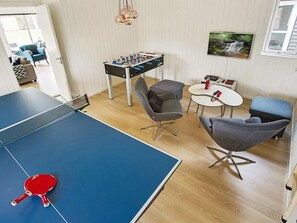 A room with a ping pong table, a foosball table, two grey armchairs, a small white table with a red bottle, and a framed picture on the wall. - 5 Star Holiday Home in Liseleje (Liseleje)