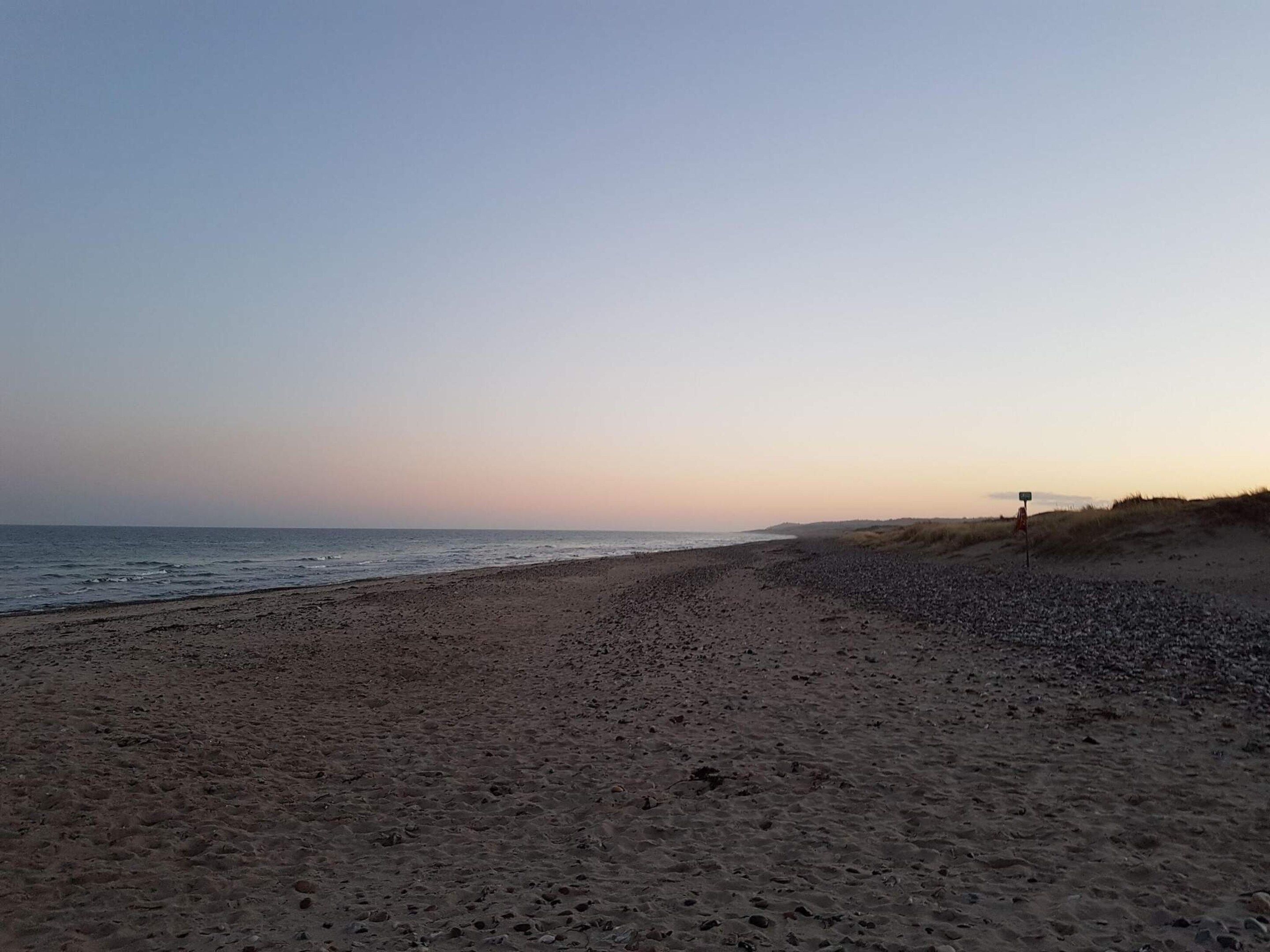 A beach with a clear sky, a sandy shore, and a dune in the background.