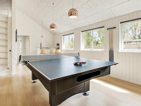 Una mesa de ping pong en una habitación con ventanas amplias, paredes de madera y lámparas colgantes.