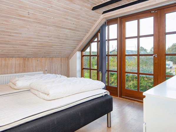 A bedroom with a sloped ceiling, a bed with white bedding, a wooden bench, and a view through large windows.