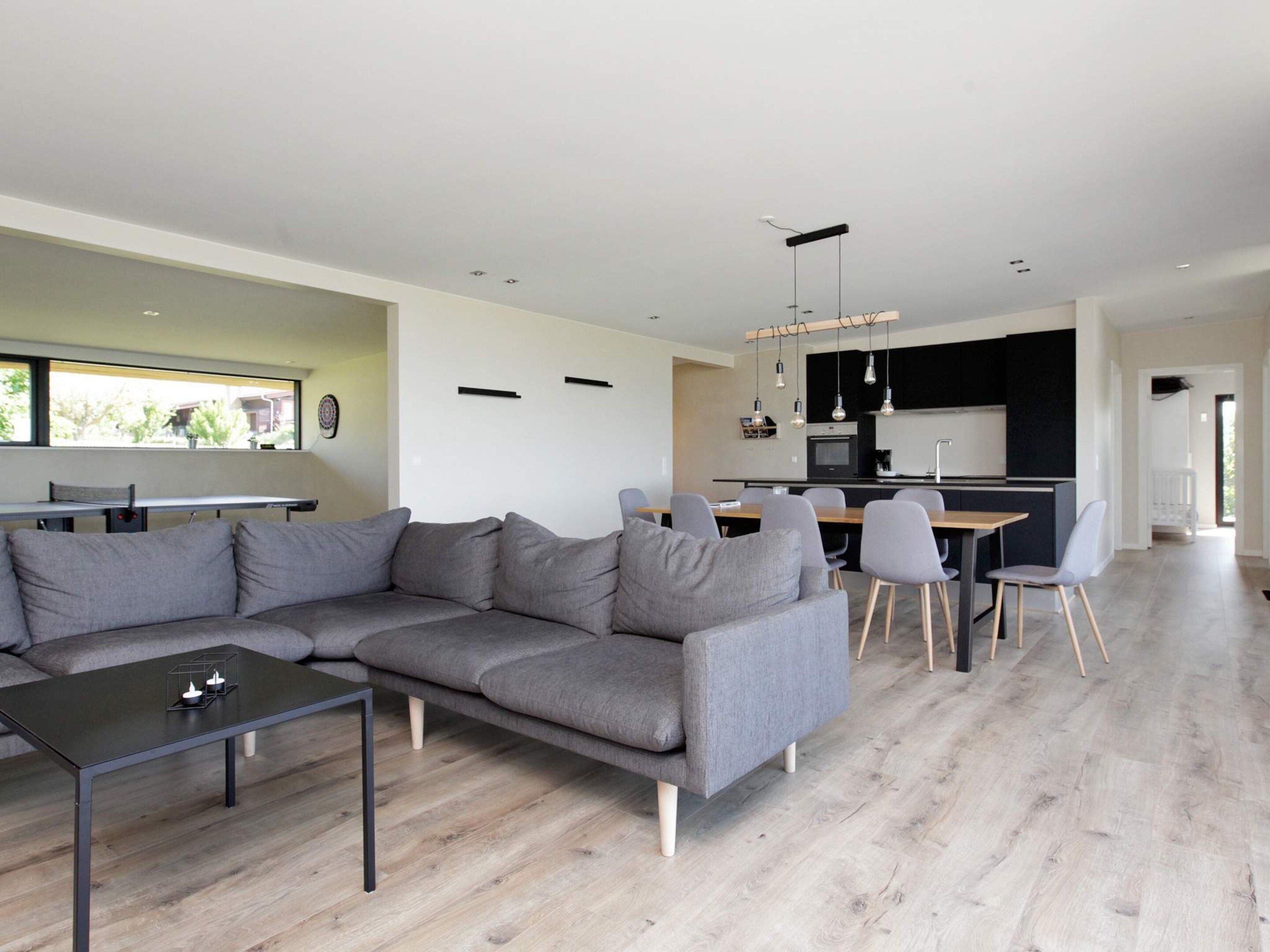 A modern living room with a grey sectional sofa, a dining table with chairs, and a kitchen area in the background.