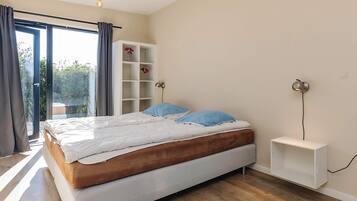 A bedroom with a bed, a white bedside table, a wall-mounted lamp, and a built-in shelving unit.