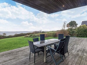 Terraza exterior con mesa y sillas, con vista a un área verde y al mar.