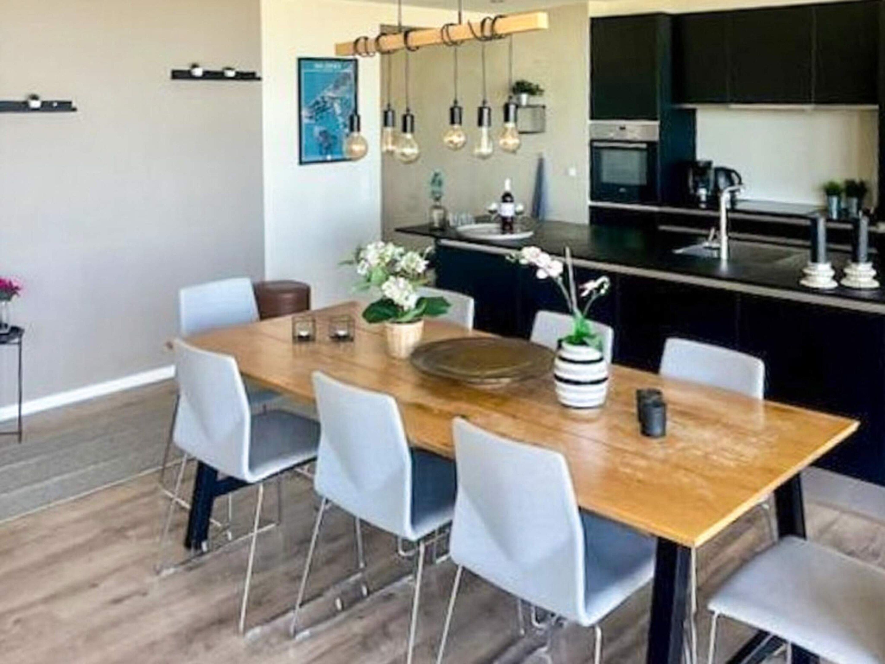 A modern kitchen with a wooden dining table, light-colored chairs, hanging pendant lights, and a built-in oven.
