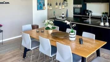 A modern kitchen with a wooden dining table, light-colored chairs, hanging pendant lights, and a built-in oven.