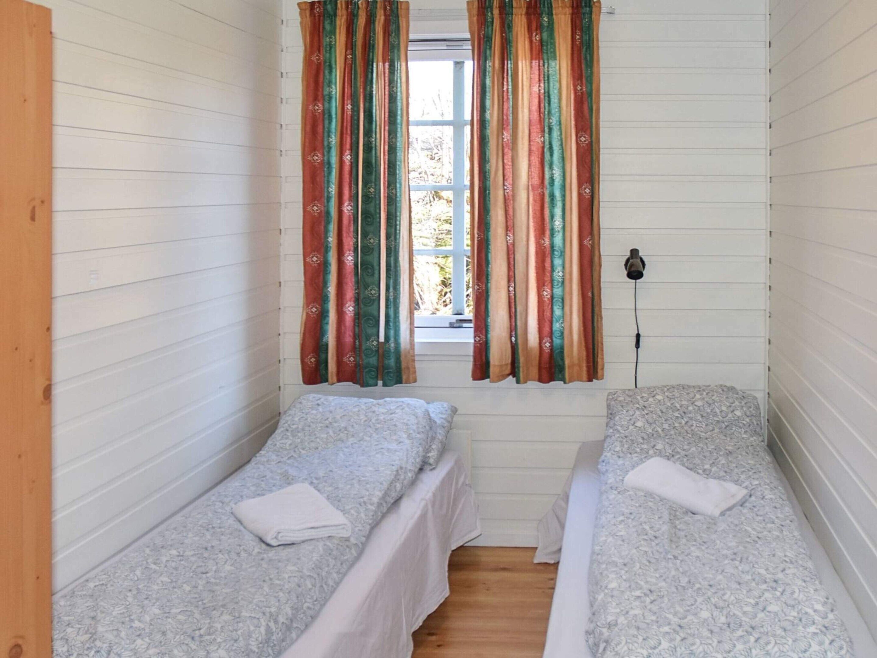 Un petit logement composé d’une seule pièce, avec deux lits simples, une fenêtre avec des rideaux et une lampe fixée au mur.