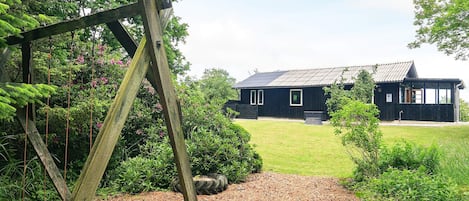 A wooden swing set in a garden leading to a black building with a metal roof.