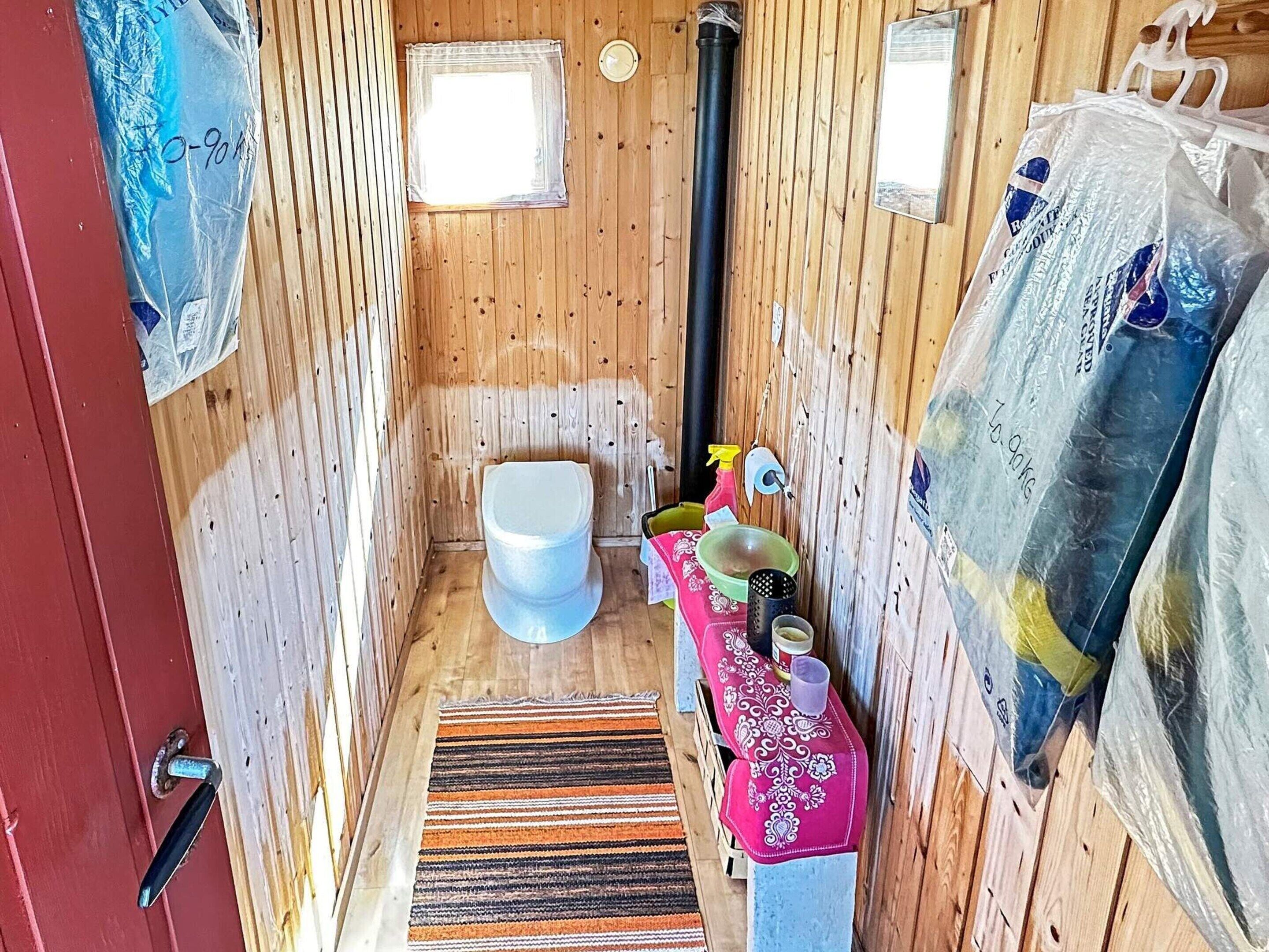 Egy kis, fából készült, egyszerű fürdőszoba, amelyben található egy WC, egy kis ablak, egy faajtó, valamint egy mosdókagyló, piros törölközővel és csíkos szőnyeggel.