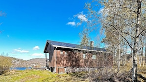 Eine rustikale Holzhütte mit geneigtem Dach, eingebettet in eine natürliche Landschaft mit einem See und Hügeln im Hintergrund.