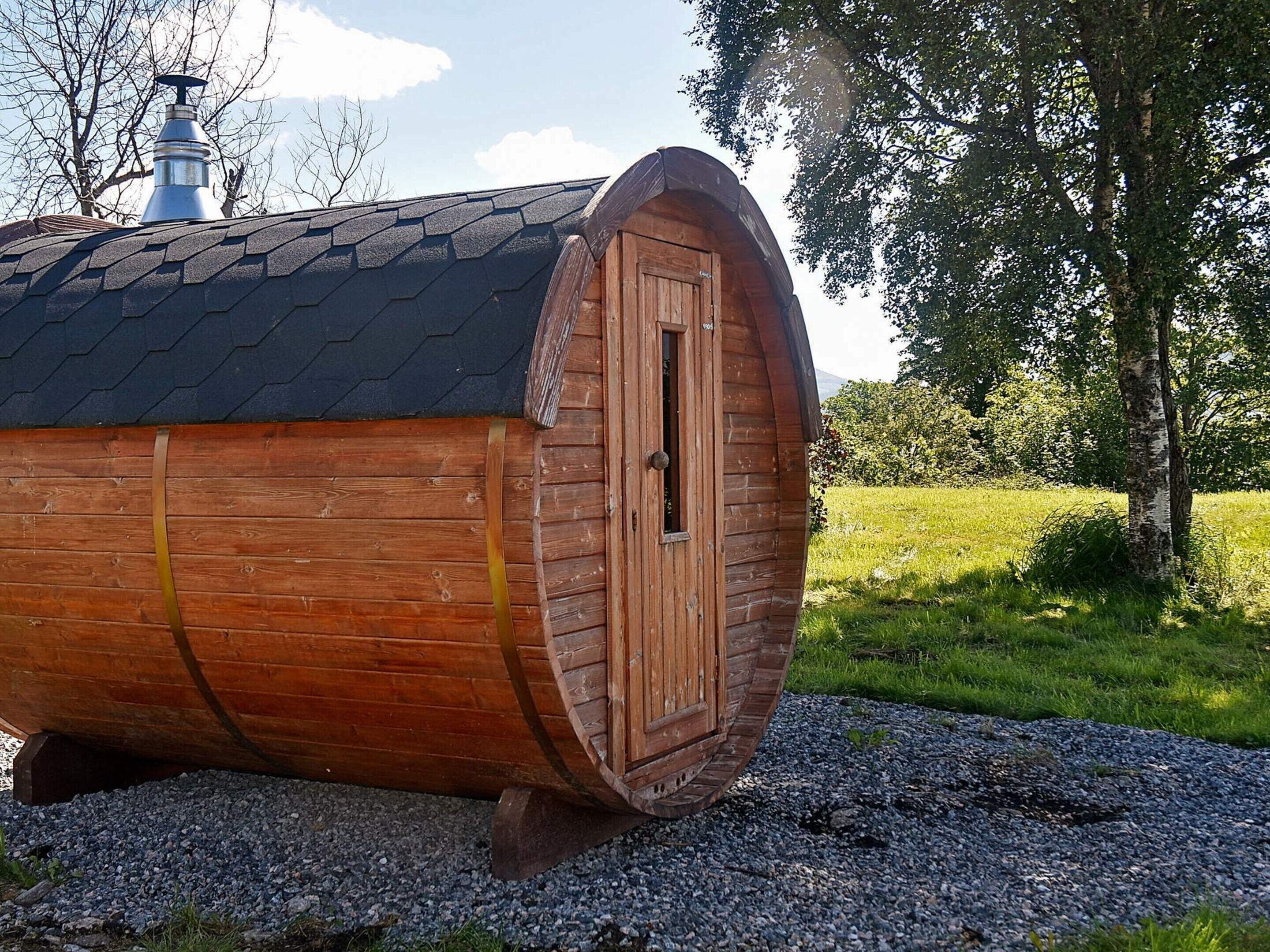 Ein bauchiges, holzverkleidetes Gebäude mit Tür und Schornstein, umgeben von Bäumen und Gras.