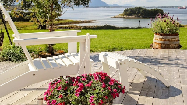 A wooden deck with a white chair, a hanging flower pot, and a view of a lake and distant mountains. - Holiday Home in Tornes I Romsdal (Hustadvika)