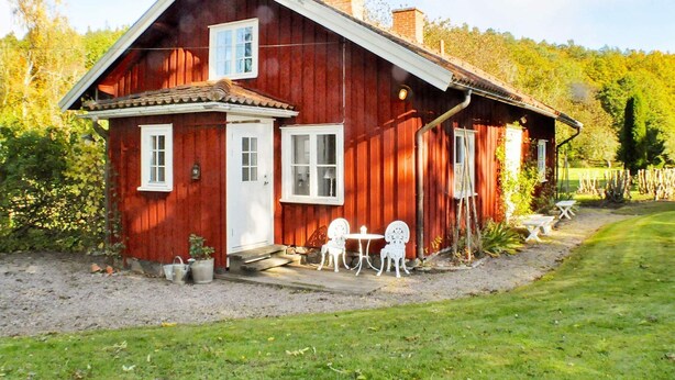 A red wooden house with a white door and windows, a chimney, and a small patio with outdoor furniture. - Holiday Home in Vargön (Västra Tunhem)