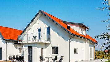 Una casa moderna de dos pisos con techo rojo, un balcón y un patio con mobiliario exterior.