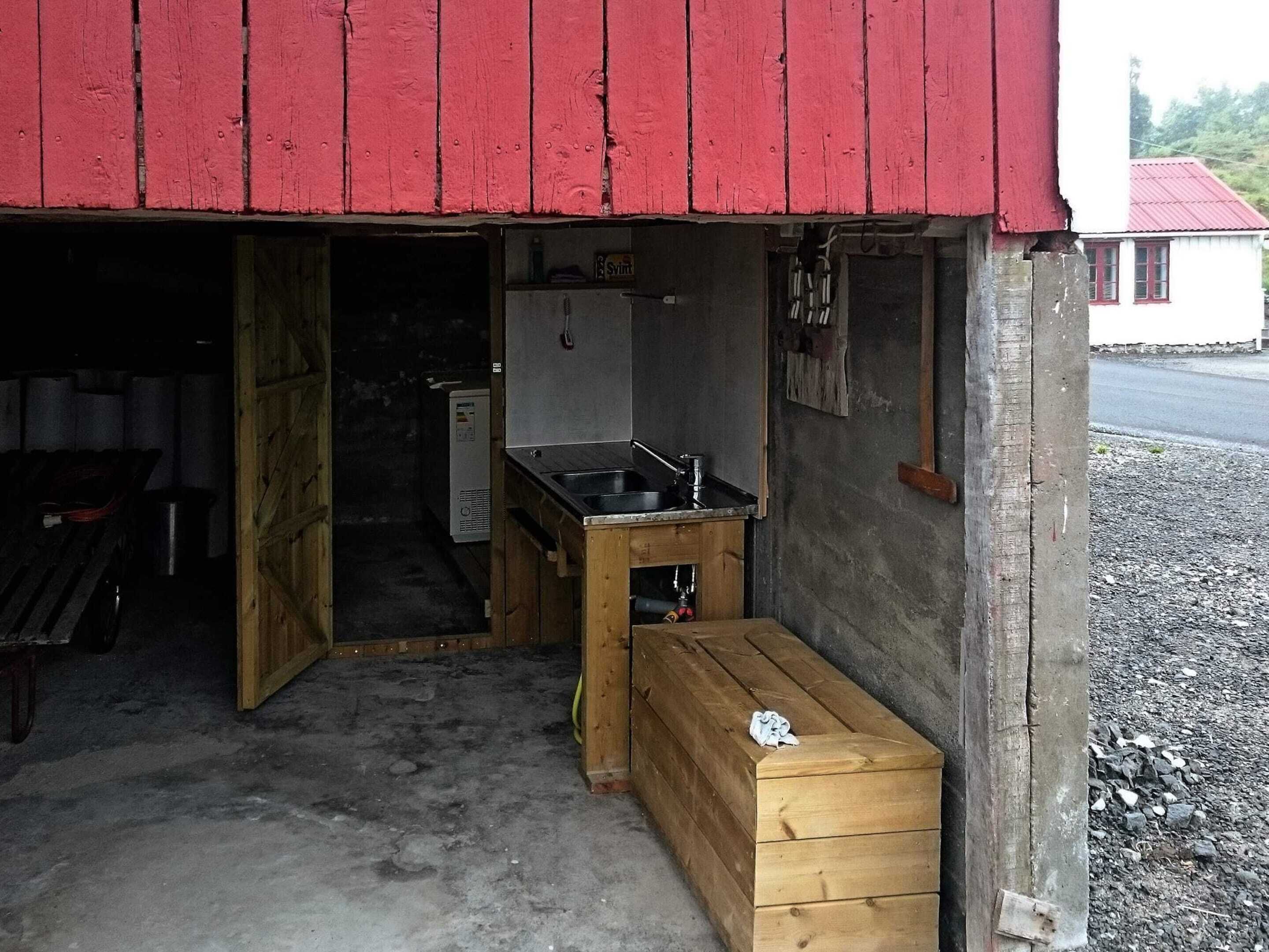 Un pequeño edificio rústico con fachada roja, un banco de madera, un lavabo y una mesita.
