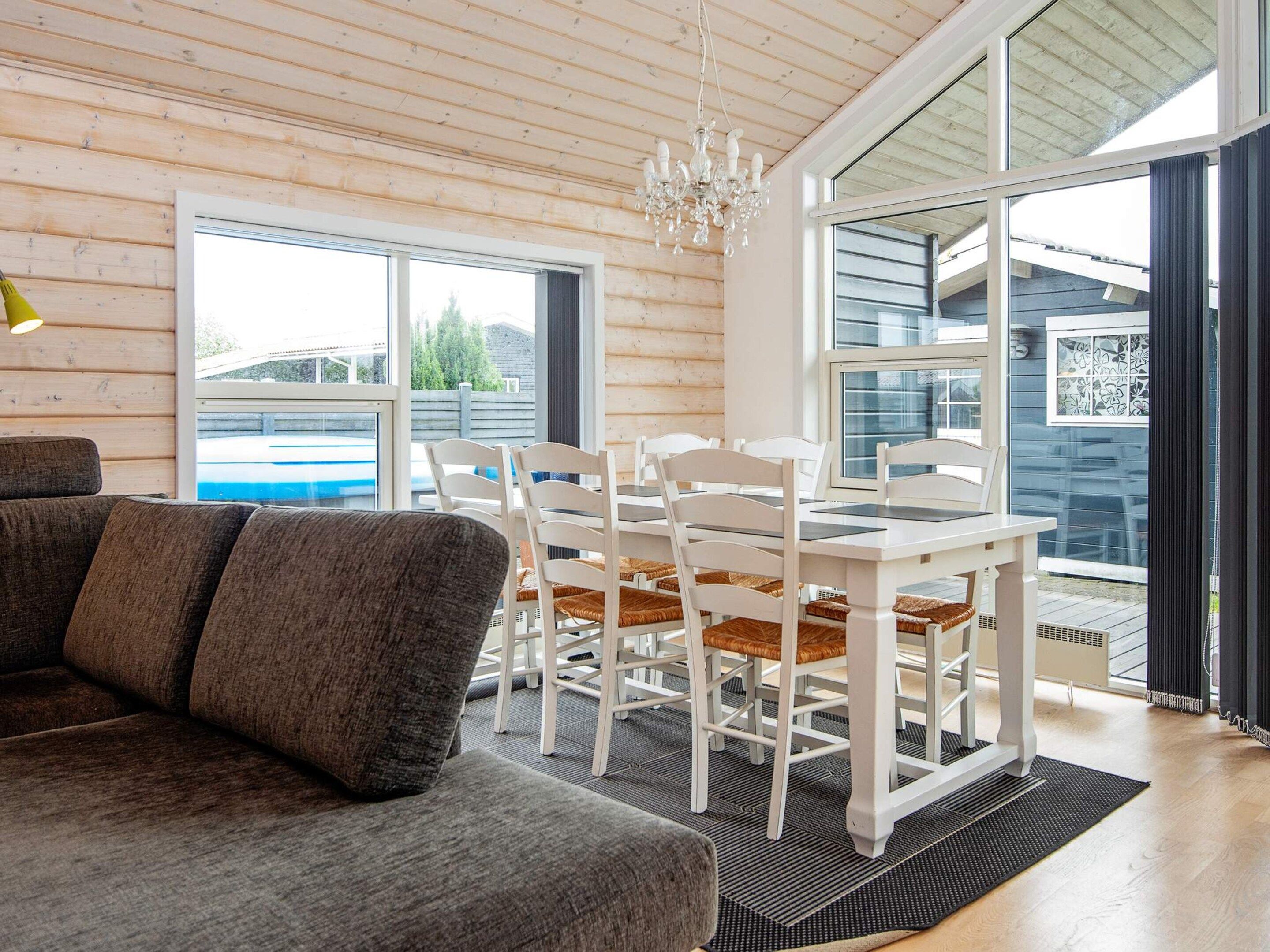 A modern interior with a wooden ceiling, a dining area with white chairs and a table, and a large window offering a view of the outdoors.