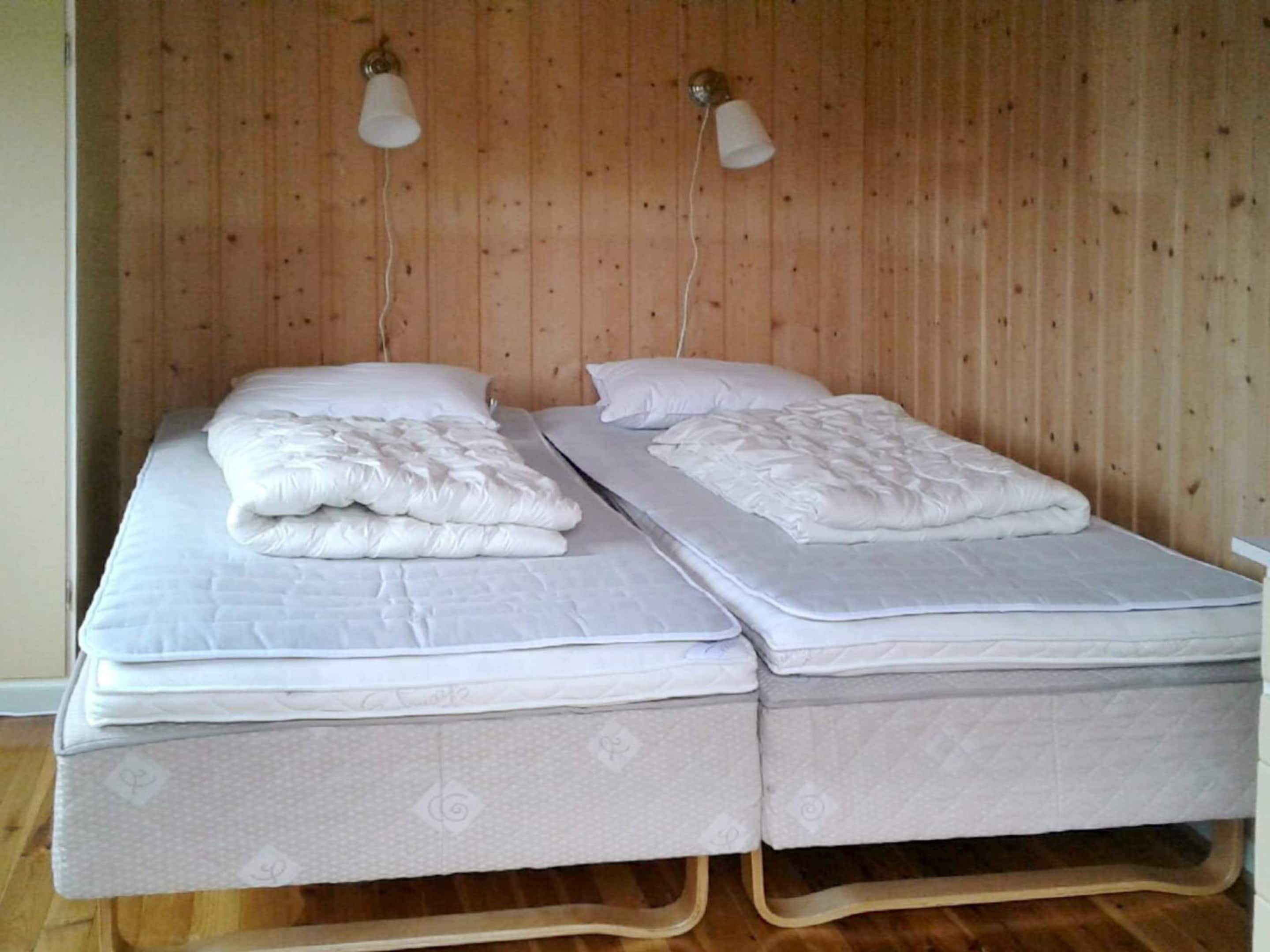 A double bed with two pillows and a blanket, wooden walls, and two wall-mounted lamps.