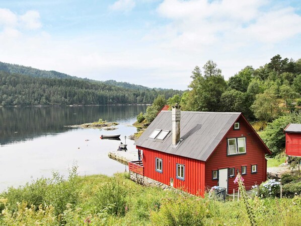 A red house with a sloped roof, a chimney, and a dock extending into a calm lake surrounded by forested hills. - 5 Person Holiday Home in Lonevag (Osteroy)