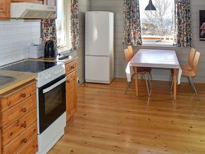 Eine kleine Küche mit Herd, Backofen und Kühlschrank, Holzschränken, einem Esstisch mit Stühlen und einem Fenster mit Vorhängen.
