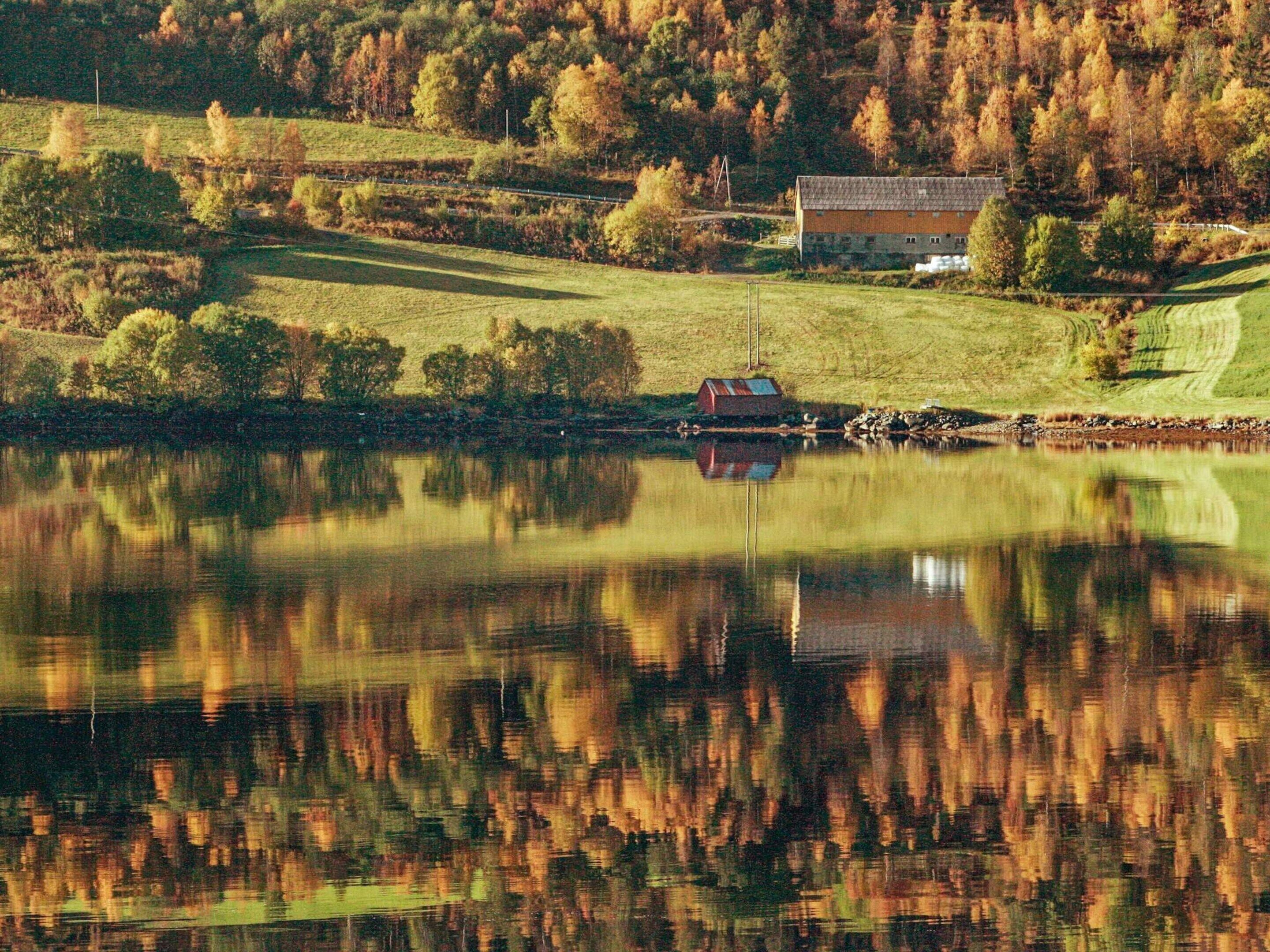A serene lakeside scene with a house and a barn, surrounded by autumn-colored trees and a calm water body reflecting the landscape.