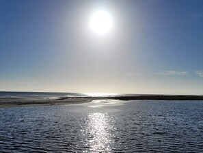 Een strand met kalm water dat zonlicht weerspiegelt, een heldere lucht en land in de verte.