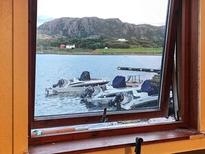 Udsigt gennem et vindue af en lysthavn med flere både i havn, en kaj der strækker sig ud i vandet, og et bjergrigt landskab i baggrunden.