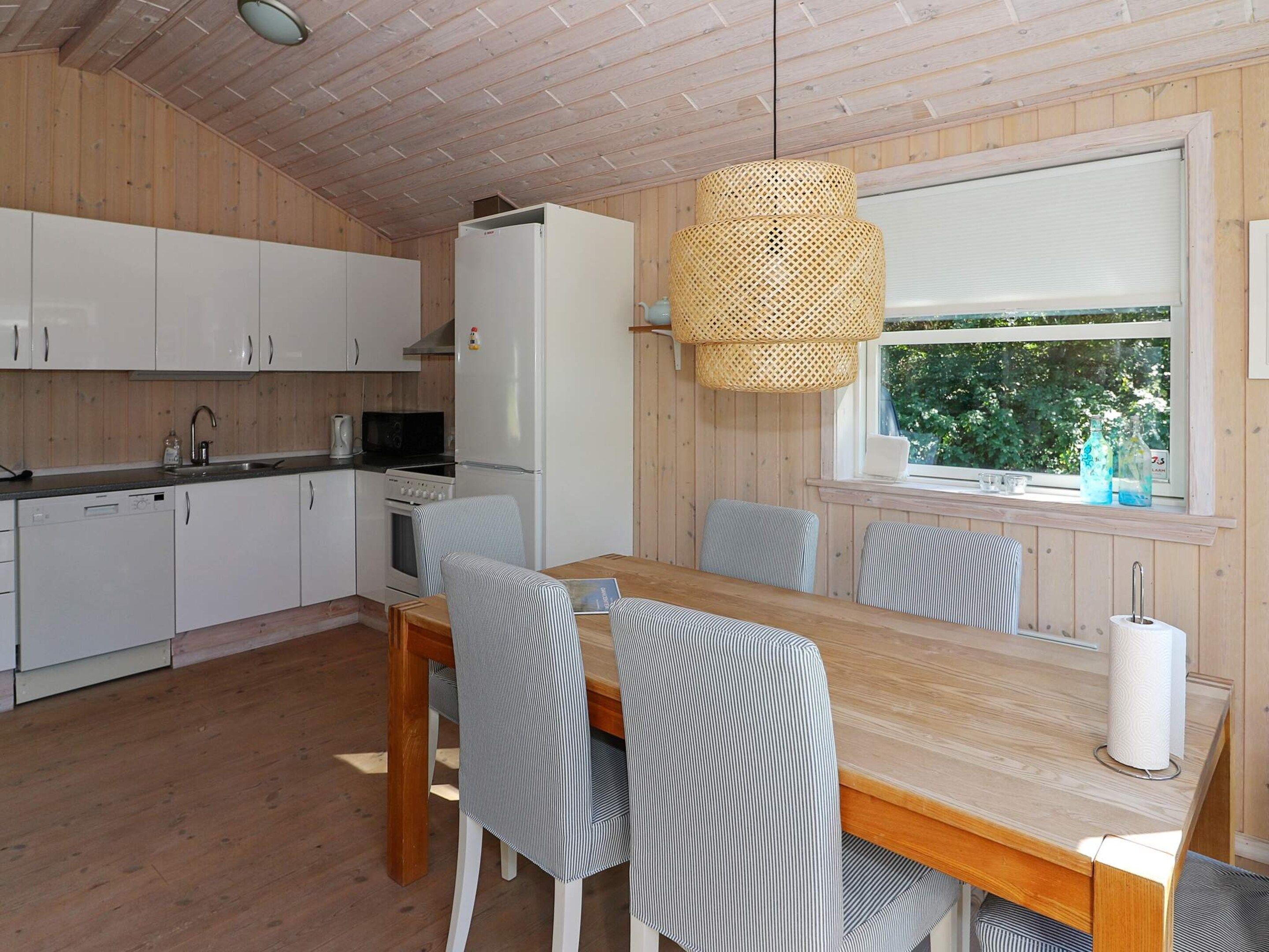 Een keuken met witte kastjes, een fornuis en een koelkast. Er staat een eettafel met stoelen en een raam met uitzicht op groen.