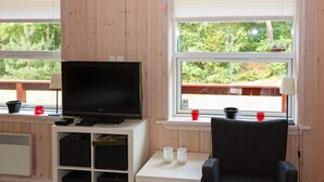 Ein Zimmer mit Fernseher auf einem weißen Schrank, einem schwarzen Sessel, einem kleinen weißen Tisch und Fenstern mit Blick ins Grüne.