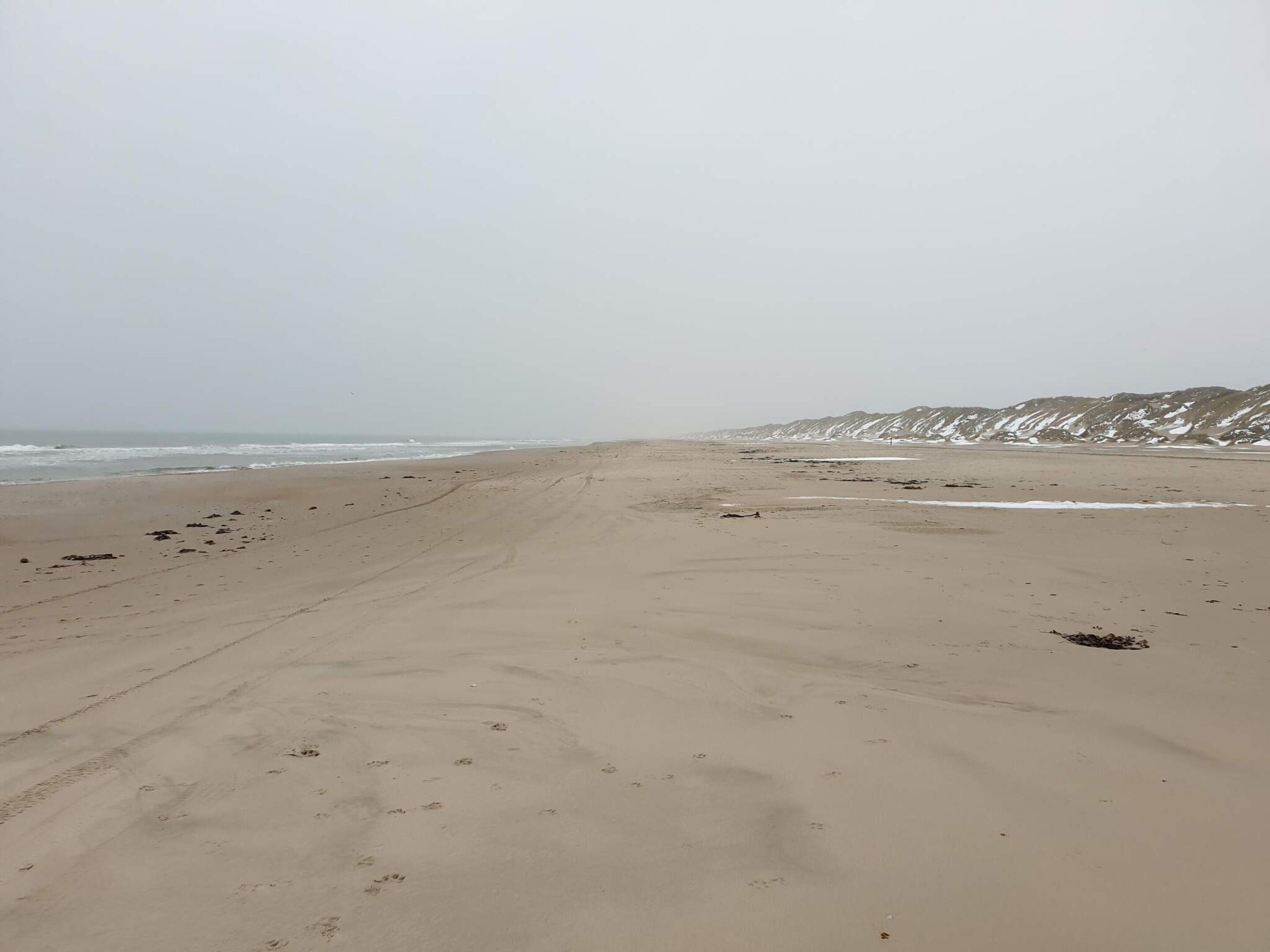 En bred sandstrand med fotspår och en avlägsen utsikt över snötäckta kullar.