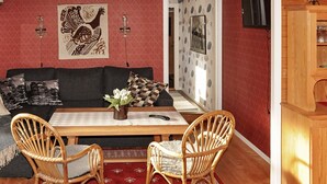Een kamer met een roodgekleurd vloerkleed, een houten tafel, twee rieten stoelen, een zwarte bank en een houten kast.