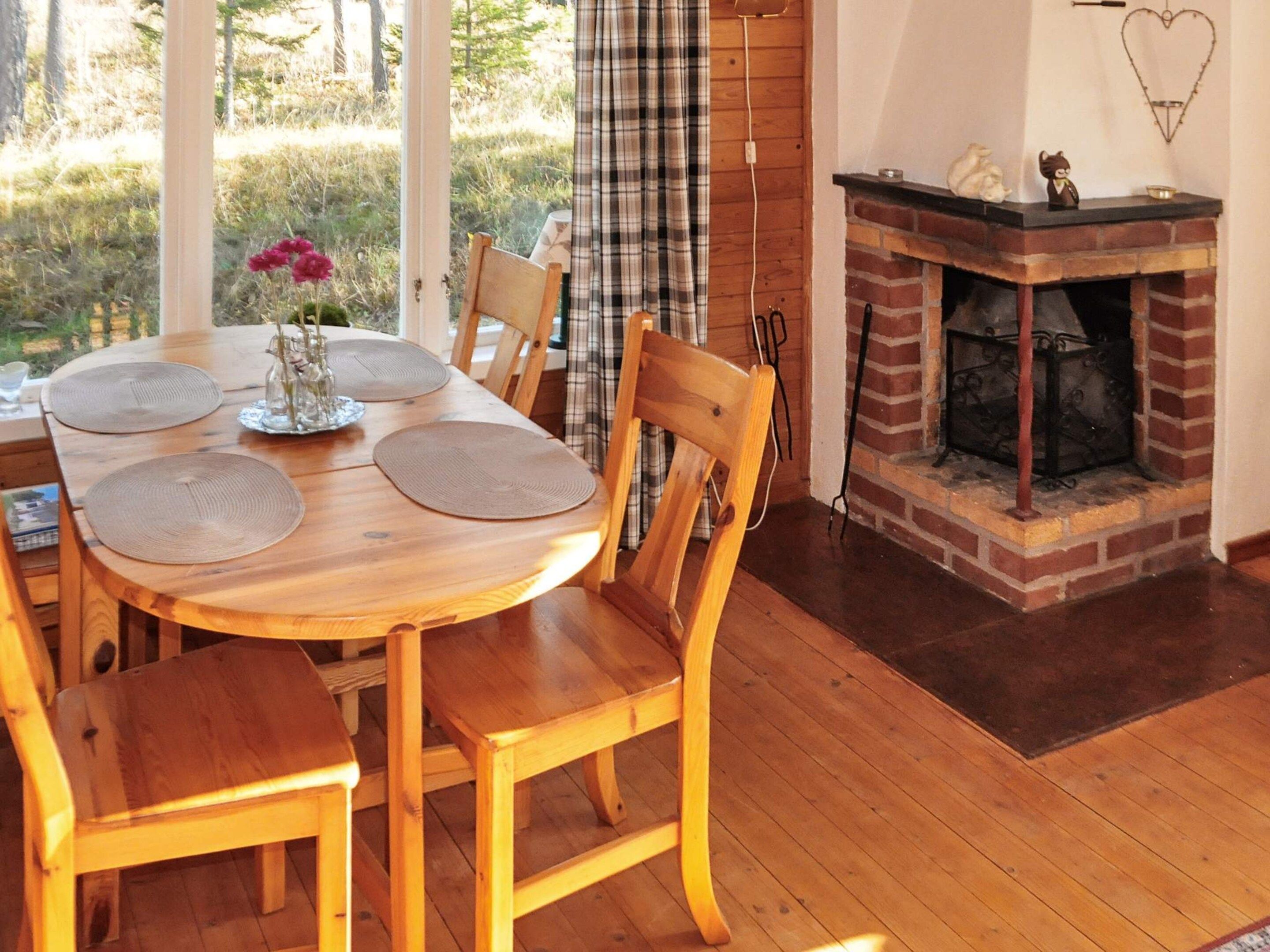 A wooden dining table with placemats, surrounded by chairs, in a room with a brick fireplace and a view of greenery outside.