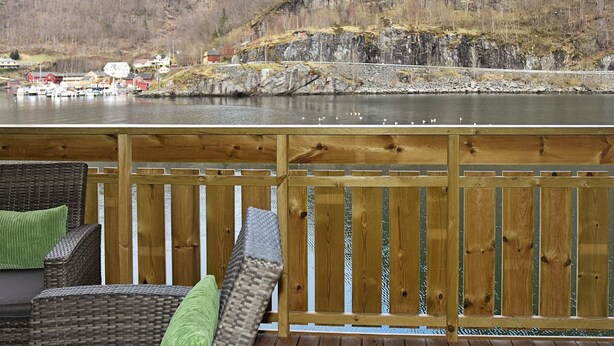 Une terrasse en bois, agrémentée de meubles et de coussins en osier, qui offre une vue sur une rivière paisible, des bateaux et un paysage montagneux.