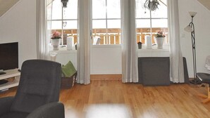 Un salon avec un plancher en bois, un fauteuil noir, un meuble pour la télévision et une fenêtre avec des rideaux.