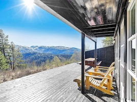 Una terraza de madera con vistas a montañas, un espacio de estar cubierto con bancos y una mesa de madera.