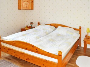 A wooden bed with a white mattress and a wooden headboard. There is a small bedside table with a lamp and a decorative item. The room has a patterned wallpaper and a wooden cabinet above the bed.