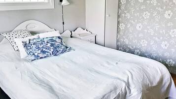 En snyggt bäddad säng med vita sängkläder och en blå, blommig prydnadskudde, ett sängbord med en lampa och en väggtapet med blommönster.
