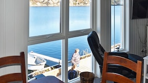 Huoneessa on suuret ikkunat, joista avautuu näkymä vesistöön. Siellä on myös puinen pöytä, musta tuoli ja seinälle asennettu televisio.