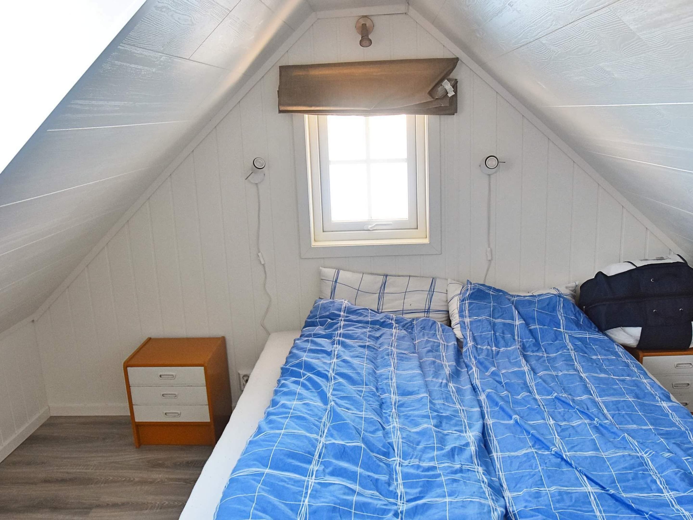 A small, sloped-roofed room with a single bed, a nightstand, a wooden drawer unit, and a window with a curtain.