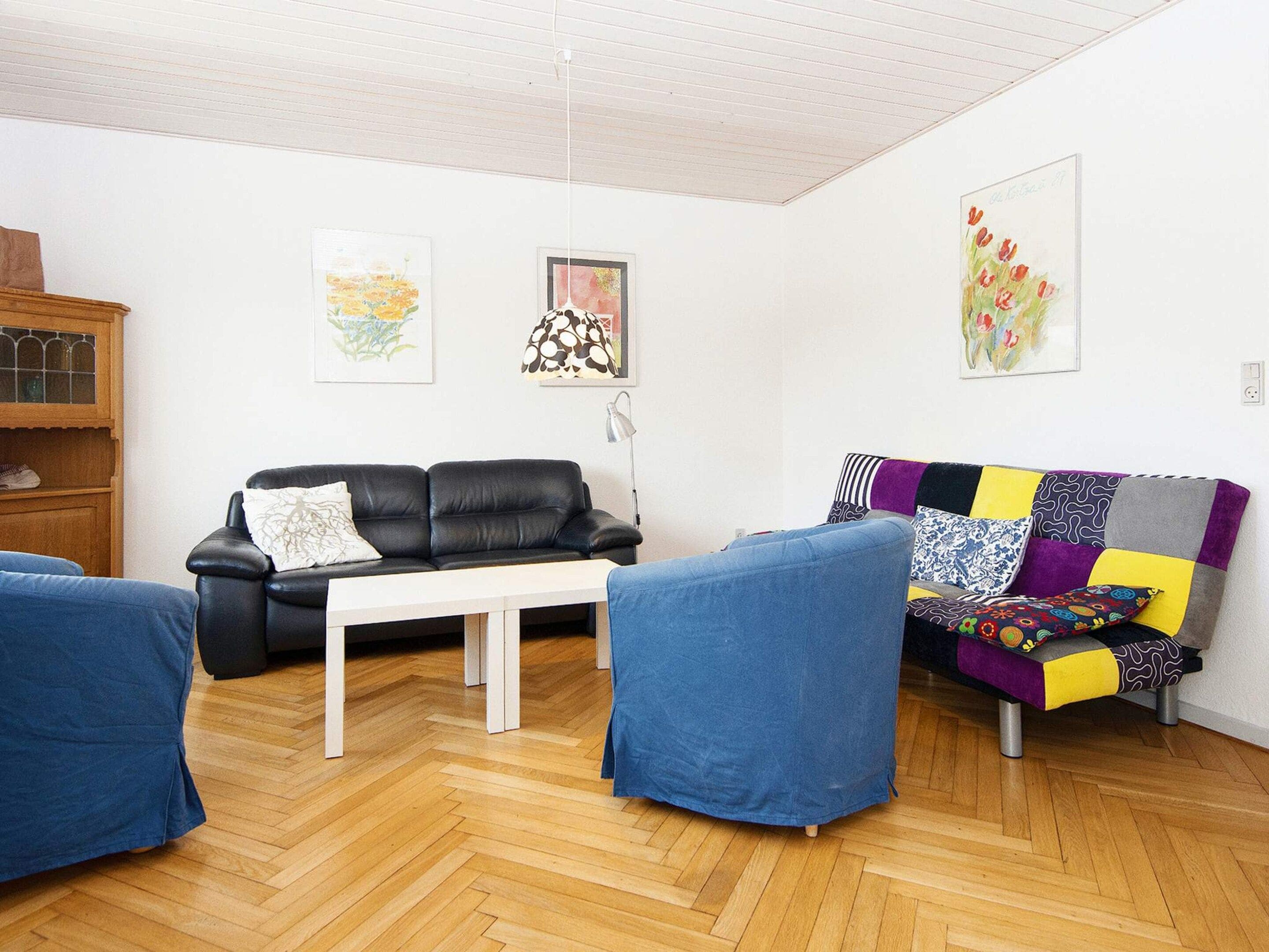 A modern living room with a black leather sofa, a white coffee table, and a colorful patchwork sofa. There are two blue armchairs and a wooden cabinet with glass doors.