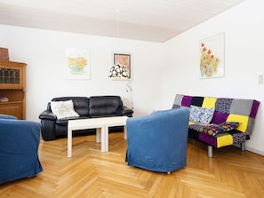 A modern living room with a black leather sofa, a white coffee table, and a colorful patchwork sofa. There are two blue armchairs and a wooden cabinet with glass doors.