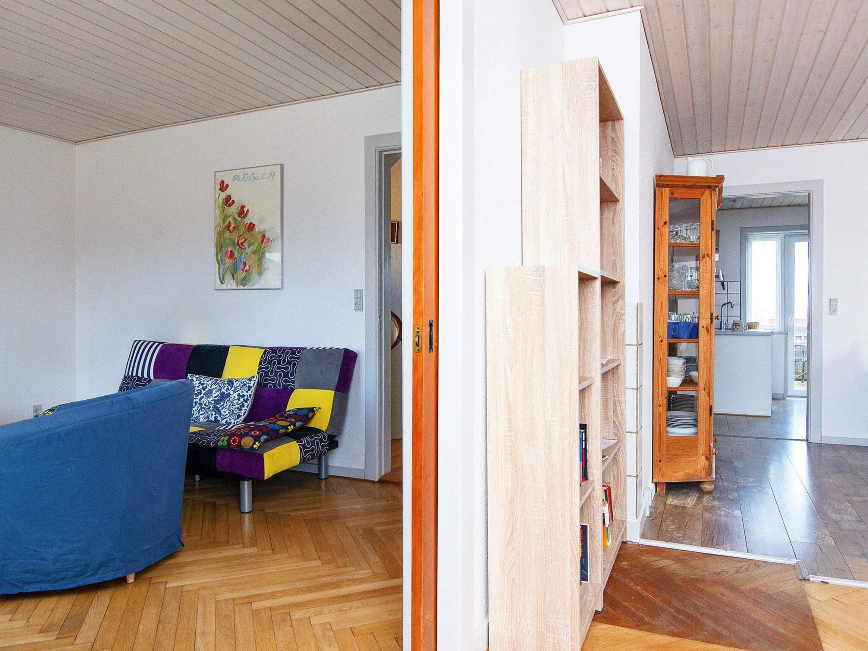 A room with a wooden floor, a blue sofa with patterned cushions, a painting on the wall, and a wooden bookshelf.