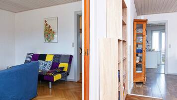 A room with a wooden floor, a blue sofa with patterned cushions, a painting on the wall, and a wooden bookshelf.
