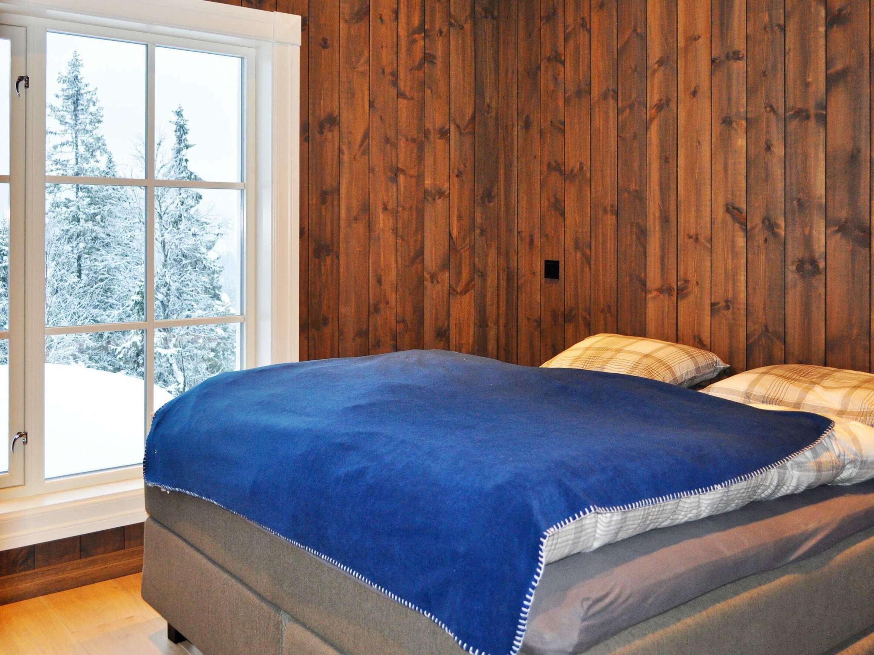Ein Schlafzimmer mit einer holzgetäfelten Wand, einem Bett mit blauer Decke und einem Fenster mit Blick auf Schnee.