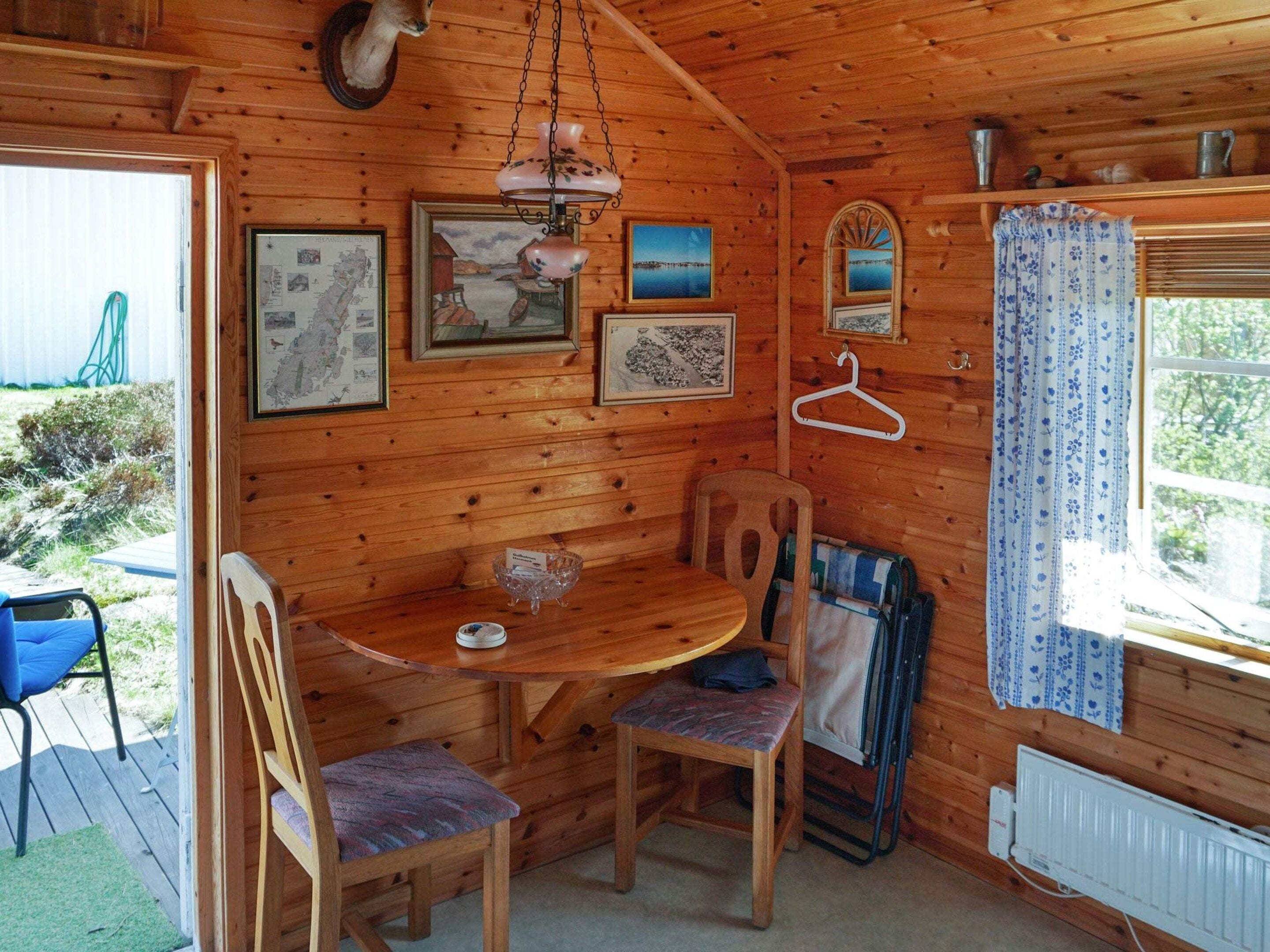A wooden cabin interior with a dining table, chairs, a hanging lamp, framed pictures, and a window with curtains.