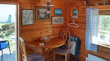 A wooden cabin interior with a dining table, chairs, a hanging lamp, framed pictures, and a window with curtains.