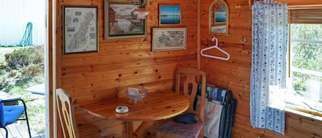 A wooden cabin interior with a dining table, chairs, a hanging lamp, framed pictures, and a window with curtains.