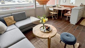 Ein modernes Wohnzimmer mit einem grauen Sofa, einem runden Holztisch mit Blumen, einer hängenden orangefarbenen Lampe und Blick auf die Straße durch große Fenster.