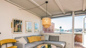 Ein modernes Wohnzimmer mit einem grauen Ecksofa, einem gelben Sessel und einem runden Couchtisch. An der Wand hängen gerahmte Bilder und ein großes Fenster bietet einen Ausblick nach draußen.