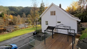 A white building with a sloped roof, a covered wooden deck, and a view of a river and forested hills. - 8 Person Holiday Home in Lindesnes (Lindesnes)
